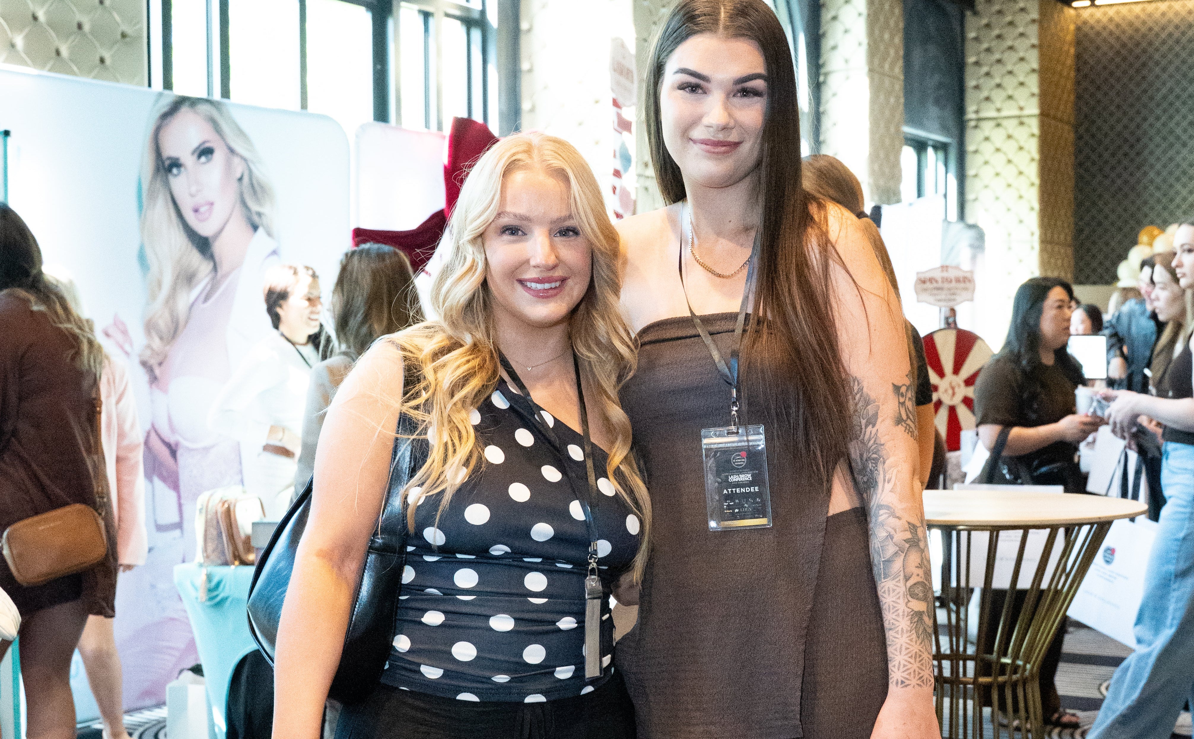 Two women posing together in a stylish indoor setting with a large LASHBOXLA poster in the background.